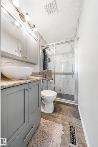 Bathroom featuring a gray vanity with a vessel sink, a shower enclosure with a sliding glass door, white rectangular wall tiles with veining, and a toilet - 2437 89 Street, Edmonton, AB 