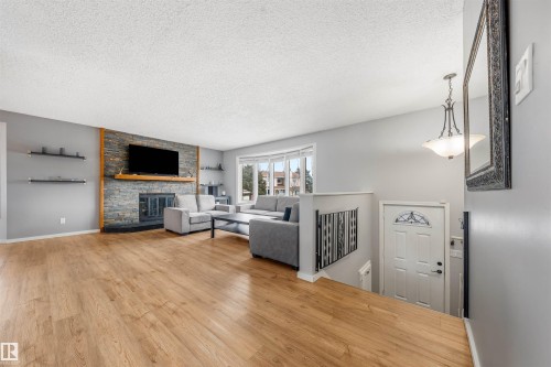 Spacious living area featuring wood-finish flooring, a stone-clad fireplace with a wood mantel, a large bay window, and a decorative railing leading to the lower level - 2437 89 Street, Edmonton, AB 