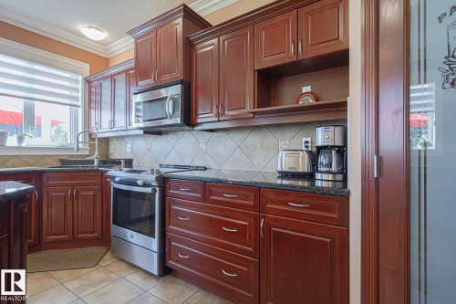Kitchen featuring rich wood cabinetry, stainless steel appliances, dark stone countertops, tile backsplash, and tile flooring - 10731 150 Street Nw, Edmonton, AB - Indoor Photo Showing Kitchen
