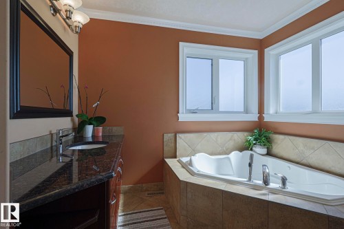 Bathroom featuring a built-in jetted tub with tile surround, dual windows, and a vanity with a dark countertop, undermount sink, and chrome faucet - 10731 150 Street Nw, Edmonton, AB - Indoor Photo Showing Bathroom