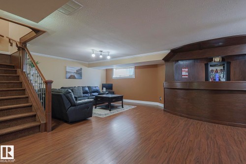 Spacious lower-level area featuring wood-finish flooring, a built-in wood-paneled bar, a carpeted staircase with wood banister and metal spindles, perimeter crown molding, and a track lighting fixture - 10731 150 Street Nw, Edmonton, AB - Indoor