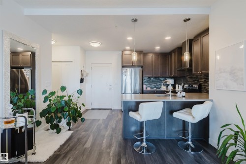 Open-concept kitchen featuring wood-finish flooring, dark wood cabinetry, a stone-look countertop with a breakfast bar, stainless steel appliances, and pendant lighting - 316 1316 Windermere Way, Edmonton, AB - Indoor Photo Showing Kitchen With Stainless Steel Kitchen With Upgraded Kitchen