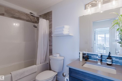 Bathroom featuring a white tub, shower with stone-look tile surround, toilet, and vanity with a gray countertop and blue base - 316 1316 Windermere Way, Edmonton, AB - Indoor Photo Showing Bathroom
