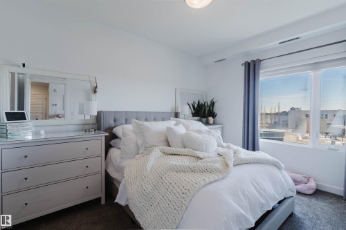 Bright bedroom featuring a large window, a tufted fabric headboard, a ceiling-mounted light fixture, carpeted flooring, and a mirrored dresser - 316 1316 Windermere Way, Edmonton, AB - Indoor Photo Showing Bedroom