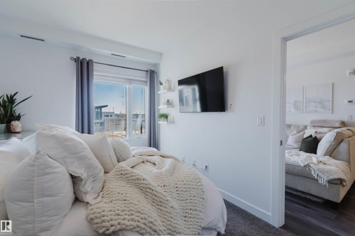 Bright interior space featuring a window with gray privacy drapes, wall-mounted shelving, and white baseboards - 316 1316 Windermere Way, Edmonton, AB - Indoor Photo Showing Bedroom
