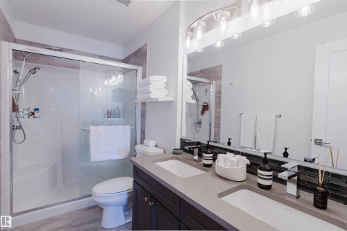 Spacious bathroom featuring a double vanity with integrated sinks, a wide mirror with overhead lighting, a glass-enclosed shower with a built-in bench, and wood-finish flooring - 316 1316 Windermere Way, Edmonton, AB - Indoor Photo Showing Bathroom