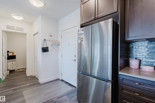 Kitchen area featuring wood-finish flooring, stainless steel refrigerator, dark wood-finish cabinetry, and a multi-toned blue tile backsplash - 316 1316 Windermere Way, Edmonton, AB - Indoor