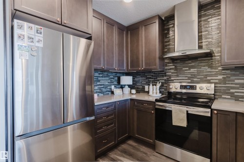 Kitchen featuring dark wood cabinetry, light-toned countertops, a mosaic tile backsplash, stainless steel appliances, and wood-finish flooring - 316 1316 Windermere Way, Edmonton, AB - Indoor Photo Showing Kitchen With Stainless Steel Kitchen With Upgraded Kitchen
