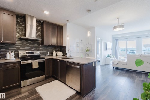 Open-concept kitchen featuring dark wood cabinetry, stainless steel appliances, a mosaic tile backsplash, light-toned countertops, and wood-finish flooring - 316 1316 Windermere Way, Edmonton, AB - Indoor Photo Showing Kitchen With Upgraded Kitchen