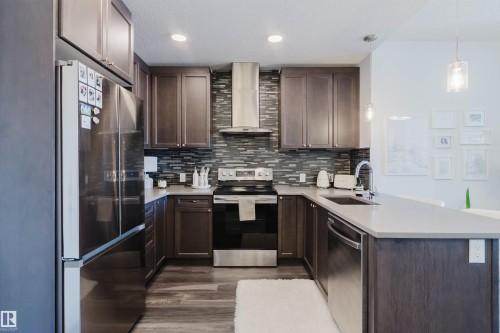 Modern kitchen featuring wood-finish cabinetry, stainless steel appliances, a mosaic tile backsplash, and light-toned countertops - 316 1316 Windermere Way, Edmonton, AB - Indoor Photo Showing Kitchen With Stainless Steel Kitchen With Upgraded Kitchen