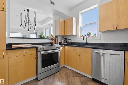 Kitchen featuring light wood cabinetry, dark countertops, stainless steel appliances, and wood-finish flooring in a herringbone pattern - 101 8108 109 Street, Edmonton, AB - Indoor Photo Showing Kitchen