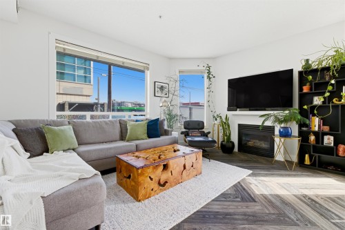 Spacious living area featuring large windows, wood-finish flooring in a herringbone pattern, a built-in fireplace with a white mantel, and ample recessed lighting - 101 8108 109 Street, Edmonton, AB - Indoor Photo Showing Living Room With Fireplace