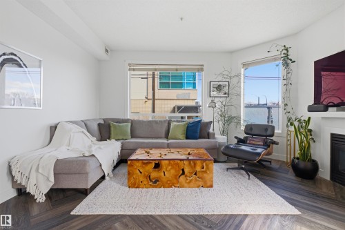 Living area featuring large windows, wood-finish flooring in a herringbone pattern, a built-in electric fireplace with a white mantel, and recessed ceiling lighting - 101 8108 109 Street, Edmonton, AB - Indoor Photo Showing Living Room With Fireplace