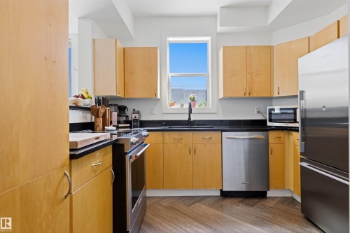 Kitchen featuring wood-finish cabinetry, dark countertops, stainless steel appliances, a stainless steel sink with a matte black faucet, and wood-finish flooring in a herringbone pattern - 101 8108 109 Street, Edmonton, AB - Indoor Photo Showing Kitchen