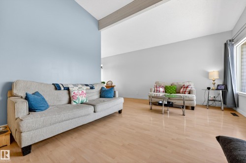 Bright living area featuring light wood-finish flooring, a vaulted ceiling with a visible beam, and a large window with curtain rod - 5304 90A Avenue, Edmonton, AB 
