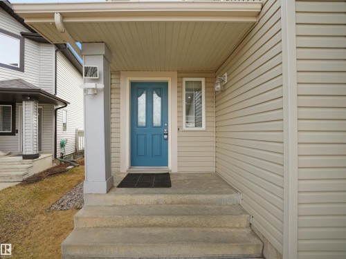 Inviting entryway featuring a teal panel door with decorative glass inserts, concrete steps, a covered porch, beige siding, and exterior lighting fixtures - 7407 Singer Landing Landing, Edmonton, AB - Outdoor With Exterior