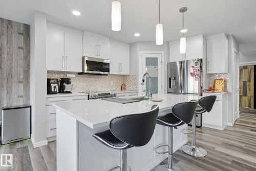 Modern kitchen featuring a central island with a solid surface countertop, white cabinetry, integrated stainless steel appliances, a mosaic tile backsplash, and wood-finish flooring - 2039 Wonnacott Way, Edmonton, AB 