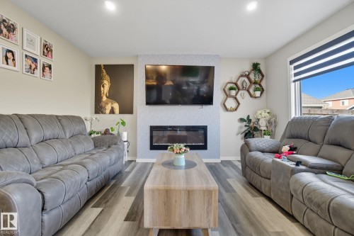 Living area featuring wood-finish flooring, a wall-mounted electric fireplace with a textured surround, recessed lighting, a large window with horizontal blinds, and a built-in wall shelf system - 2039 Wonnacott Way, Edmonton, AB 