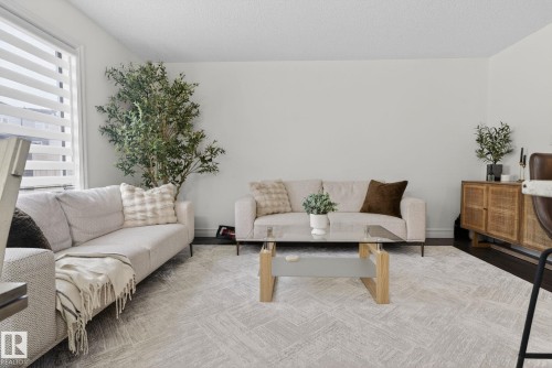 Living area featuring light-colored walls, a large window with horizontal blinds, and dark flooring partially covered by a patterned area rug - 1014 162 St Sw, Edmonton, AB - Indoor Photo Showing Living Room