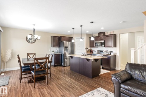 Open-concept kitchen and dining area featuring wood-finish flooring, dark wood cabinetry, stainless steel appliances, a kitchen island with pendant lighting, and a tiled backsplash - 6864 Evans Wynd, Edmonton, AB - Indoor