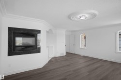 Interior living space featuring a dark-toned wood-finish flooring, a wall-mounted fireplace with a dark stone-look surround, white crown molding, arched windows, and a decorative ceiling medallion with an attached flush-mount light fixture - 