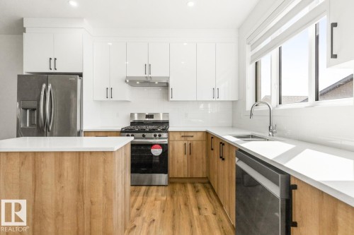 Modern kitchen featuring white upper cabinetry, wood-finish lower cabinetry, and a matching island with a light-colored countertop - 6625 46 Avenue, Beaumont, AB - Indoor Photo Showing Kitchen With Double Sink With Upgraded Kitchen