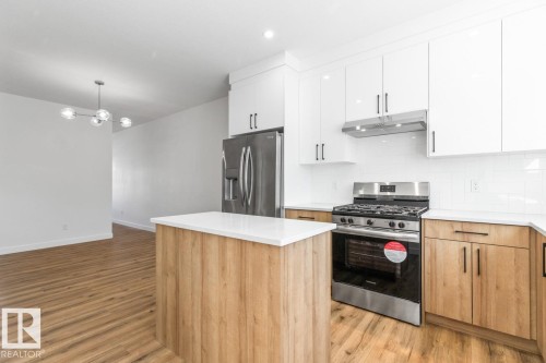 Modern kitchen featuring a central island with a solid surface countertop, stainless steel appliances including a gas range and refrigerator, white shaker-style upper cabinetry, wood-finish lower cabinetry, and wood-finish flooring - 6625 46 Avenue, Beaumont, AB - Indoor Photo Showing Kitchen