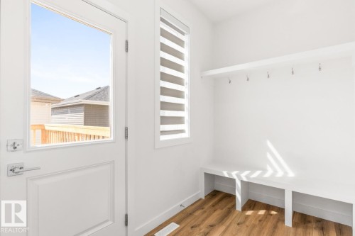 Bright mudroom space featuring a full-light exterior door with satin chrome hardware, a smaller window with privacy blinds, wood-finish flooring, a built-in bench, and a built-in shelf with hooks - 6625 46 Avenue, Beaumont, AB - Indoor Photo Showing Other Room