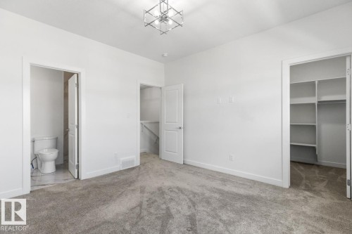 Spacious room featuring plush gray carpeting, crisp white walls, a modern geometric ceiling light fixture, a walk-in closet with built-in shelving, and an en-suite bathroom with a toilet and marble-finish flooring - 6625 46 Avenue, Beaumont, AB - Indoor