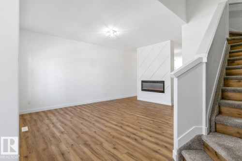 Open-concept living space with wood-finish flooring, a contemporary electric fireplace with a diagonal shiplap accent wall, a white painted staircase with carpeted treads, and a flush-mount ceiling light fixture - 6625 46 Avenue, Beaumont, AB - Indoor Photo Showing Other Room