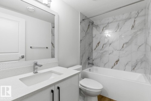 Bathroom featuring a white vanity with an integrated sink and chrome faucet, a coordinating framed mirror, and a white bathtub with a tile surround in a marble-look finish - 6625 46 Avenue, Beaumont, AB - Indoor Photo Showing Bathroom