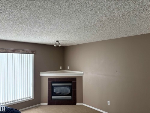 Living area featuring a corner fireplace with a tiled surround, a large window with vertical blinds, and a ceiling light fixture - 11904 18 Avenue, Edmonton, AB - Indoor Photo Showing Living Room With Fireplace