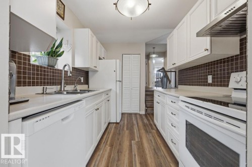 Galley kitchen featuring white cabinetry, wood-finish flooring, a mosaic tile backsplash, and a double basin stainless steel sink - 7422 188 Street, Edmonton, AB - Indoor Photo Showing Kitchen With Double Sink