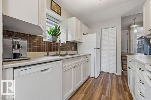 Kitchen featuring white cabinetry, dark brown square tile backsplash, wood-finish flooring, white appliances, and a double basin stainless steel sink - 7422 188 Street, Edmonton, AB - Indoor Photo Showing Kitchen With Double Sink