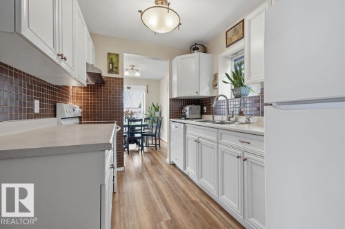 Bright kitchen featuring white cabinetry with brushed bronze hardware, a brown tile backsplash, light-toned countertops, wood-finish flooring, and a stainless steel sink with a high-arc faucet - 7422 188 Street, Edmonton, AB - Indoor Photo Showing Kitchen