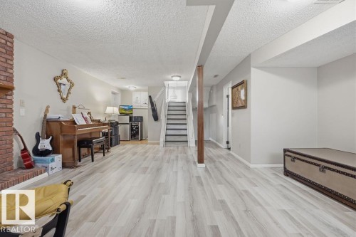 Spacious lower level featuring wood-finish flooring, a brick fireplace with a wood mantel, a staircase with dark treads, light gray wall paint, and exposed wood support beams - 7422 188 Street, Edmonton, AB - Indoor
