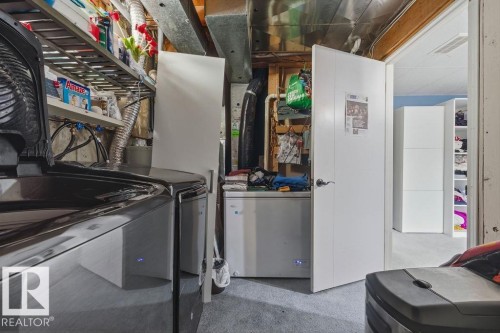 Dedicated laundry area featuring a dark-finish top-load washing machine, exposed overhead utility lines, and an adjacent white door providing access to additional finished space - 7422 188 Street, Edmonton, AB - Indoor