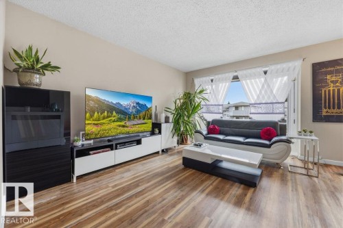 Living area featuring wood-finish flooring, a large window with sheers, light-toned walls, a textured ceiling, and an entertainment console - 7422 188 Street, Edmonton, AB - Indoor Photo Showing Living Room