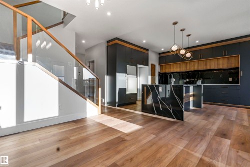 Open-concept living area featuring wide-plank wood-finish flooring, a contemporary kitchen with dark cabinetry and wood-finish accents, a waterfall island with a black and white veined surface, a black backsplash, and a modern staircase with glass railing - 372 Creekside Green, Leduc, AB - Indoor