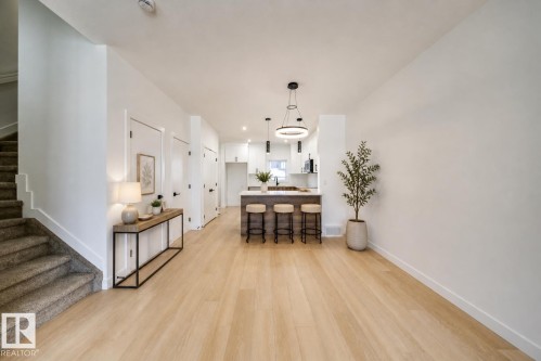 Open-concept living area featuring light wood-finish flooring, a kitchen island with seating, pendant lighting, and a staircase with carpeted treads - 86 Blackbird Bend, Fort Saskatchewan, AB - Indoor Photo Showing Other Room