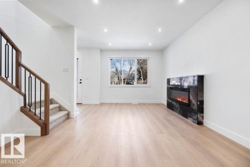 The interior features light-colored flooring, a staircase with a wooden handrail and dark balusters, and a contemporary fireplace with a dark, veined surround - 12307 91 St Nw, Edmonton, AB - Indoor Photo Showing Other Room With Fireplace