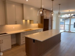 Spacious kitchen featuring an extended island with a light-toned countertop, sleek overhead cabinetry, wood-finish flooring, and under-cabinet lighting - 
