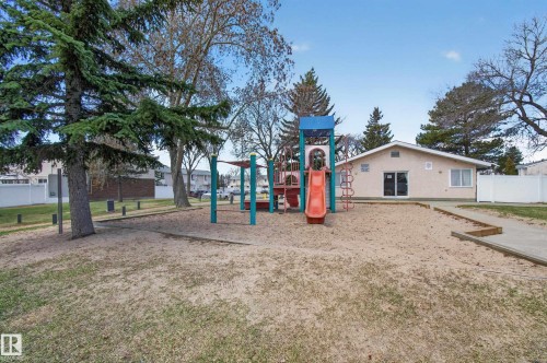 Expansive outdoor play area featuring a multi-level playset with slide, sand base, mature trees, and a beige stucco building with large windows and double doors - 18 Lakewood Village, Edmonton, AB - Outdoor