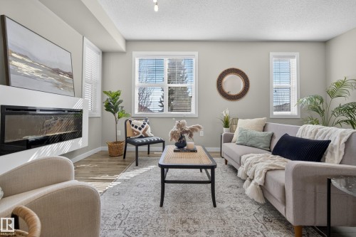 Living area featuring wood-finish flooring, a wall-mounted electric fireplace, two windows with blinds, and recessed lighting - 61 2803 James Mowatt Trail, Edmonton, AB - Indoor Photo Showing Living Room With Fireplace