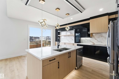 Modern kitchen featuring a central island with an undermount sink, white countertops, black high-gloss cabinetry, wood-finish upper cabinetry, and a marble-patterned backsplash - 417 Pine Point(E), Leduc, AB - Indoor Photo Showing Kitchen With Double Sink With Upgraded Kitchen