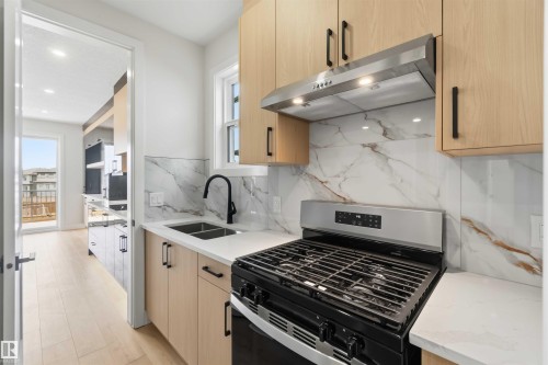 Modern kitchen featuring light wood-finish cabinetry, a stainless steel range with overhead ventilation, a dual basin sink with a matte black faucet, and a patterned backsplash with white countertops - 417 Pine Point(E), Leduc, AB - Indoor Photo Showing Kitchen With Double Sink With Upgraded Kitchen