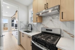 Modern kitchen featuring light wood-finish cabinetry, a stainless steel range with overhead ventilation, a dual basin sink with a matte black faucet, and a patterned backsplash with white countertops - 