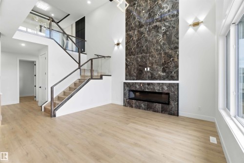 Two-story living area featuring wood-finish flooring, a contemporary linear fireplace with a dark marble-patterned surround, and a modern staircase with glass panel railings - 417 Pine Point(E), Leduc, AB - Indoor With Fireplace