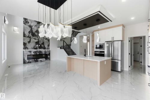 Open-concept interior featuring large format marble-look tile flooring, a modern kitchen island with a built-in sink, stainless steel appliances, a striking fireplace with a stone surround, and an elegant staircase with glass railings - 415 Pine Point(E), Leduc, AB - Indoor Photo Showing Kitchen