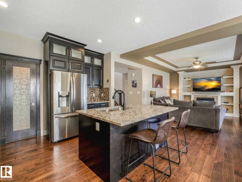 Open-concept living space with wood-finish flooring, a kitchen island with stone countertop, dark cabinetry, and integrated stainless steel appliances - 16251 135 Street Nw, Edmonton, AB - Indoor Photo Showing Kitchen With Stainless Steel Kitchen With Upgraded Kitchen